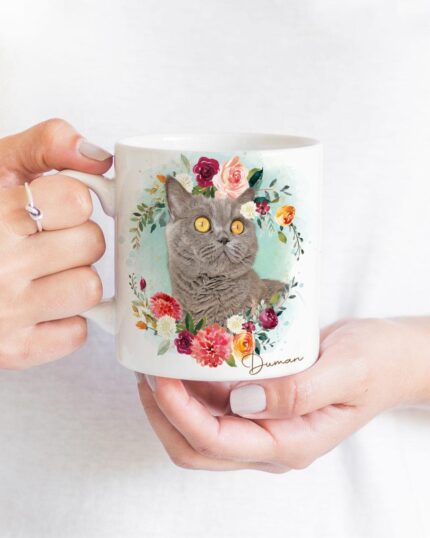 A person holds the "Flower Wreath Custom Design Mug #1," a white mug with a gray cat amid colorful flowers and greenery, "Ouman" at the bottom. The scene evokes custom pet art; the person wears a white shirt and delicate ring.