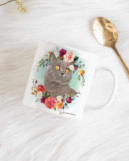The Flower Wreath Custom Design Mug #1, featuring a gray cat among vibrant flowers in a custom art style, rests on white fur beside a gold spoon and delicate flower sprigs.