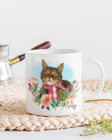 The Flower Wreath Custom Design Mug #1 features a custom pet illustration of a cat in a red plaid scarf, surrounded by colorful flowers, displayed on a woven placemat next to a gold spoon and metal coffee maker.