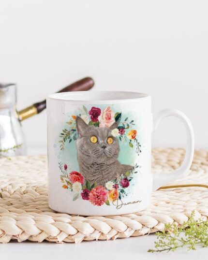 The Flower Wreath Custom Design Mug #1 features a gray cat illustration surrounded by colorful flowers on a white mug, placed on a woven placemat with a gold spoon, small green plants, and a metal coffee pot in the background.