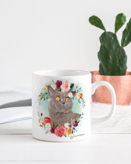 The Flower Wreath Custom Design Mug #1, featuring a photo of a gray cat, is ideal for pet lovers. It sits on a white table with a copper pot holding a cactus and an open magazine on a furry white surface in the background.