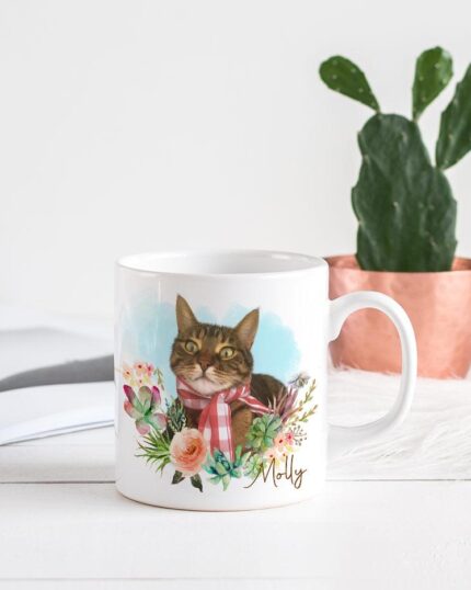 The Flower Wreath Custom Design Mug #1, featuring a cat portrait, floral accents, and the name Milly to match a custom pet canvas, rests on a white table beside an open magazine and a small cactus in a copper-colored pot.