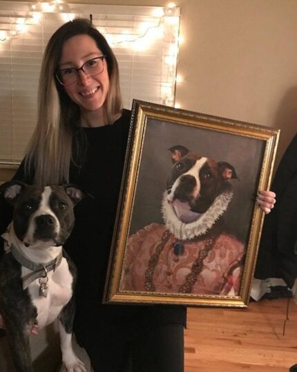 A woman smiles while holding her dog and a framed "The Noble Lady #4 - Custom Design Canvas" featuring the same dog in royal attire, with string lights and a window in the background.
