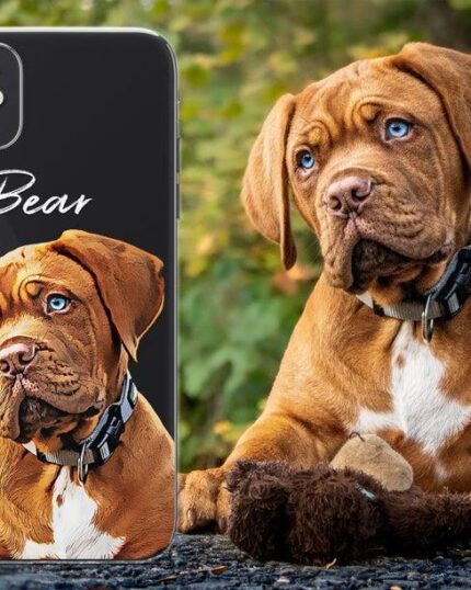 A brown puppy named Bear with blue eyes and a black collar sits outdoors beside a Personalized Transparent Phone Case featuring a custom pet canvas illustration of the puppy and "Bear" in white text, with blurred greenery in the background.