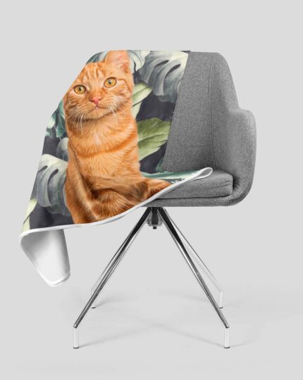 A gray modern chair with the Custom Design Pop-Art Blanket – Leaf #1 draped over it, featuring a canvas of an orange tabby cat sitting before green tropical leaves on a white background.