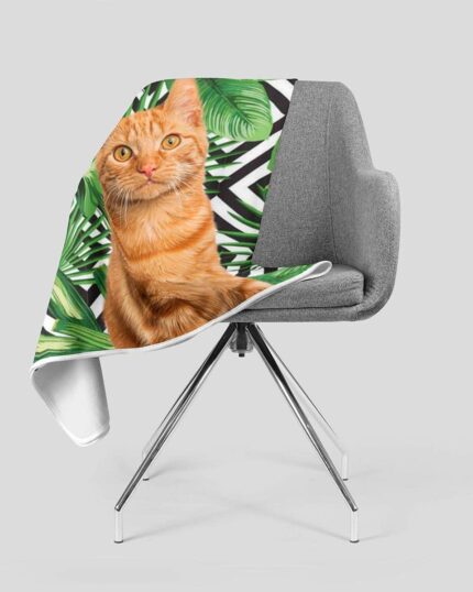 A modern gray chair is draped with the Custom Design Pop-Art Blanket – Leaf #4, featuring an orange tabby cat amid green tropical leaves and black-and-white geometric designs on a plain light gray background.