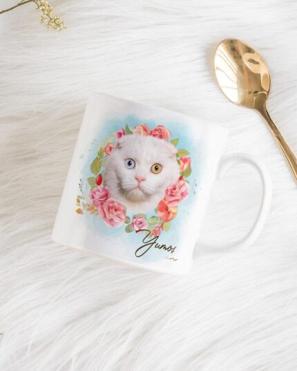 The Flower Wreath Custom Design Mug– no#7 features a colorful cat illustration with pink flowers and the word “Yours,” displayed on a white mug, placed on a furry surface beside a gold spoon and yellow flower sprigs.