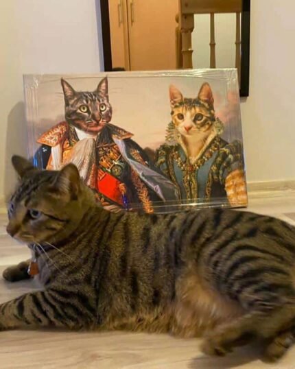 A tabby cat lies on a wooden floor in front of the "Multiple Portraits #5 King and Queen" custom pet canvas, featuring two cats dressed in royal attire. The portrait is propped against the wall near a door and mirror.