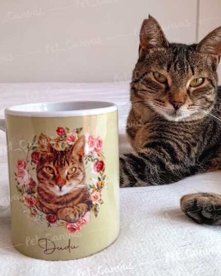 A brown tabby cat lies on a bed beside the Flower Wreath Custom Design Mug #3, which displays a canvas-style portrait of the same cat surrounded by flowers and the name "Duque" beneath the image.
