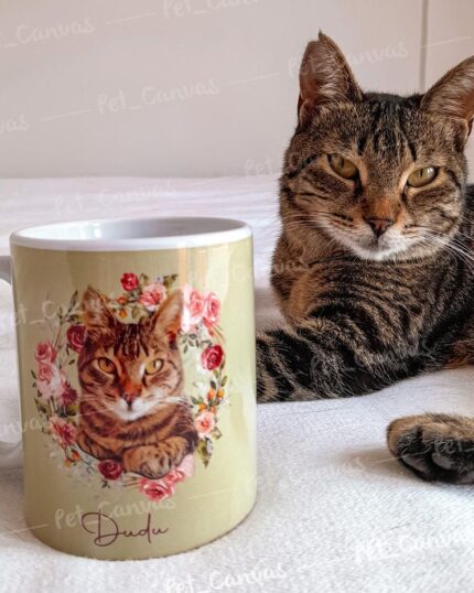 A tabby cat lies next to a mug featuring an illustrated portrait of the same cat surrounded by pink and red flowers, inspired by a custom design pet canvas. The cream-colored mug sits on a white surface.