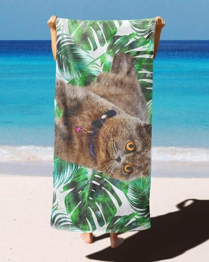 On a sandy beach, someone holds up the Custom Design 3D Printed Beach Towel – Blueberry, showcasing a gray cat and green tropical leaves with the bright blue ocean and sky in the background.