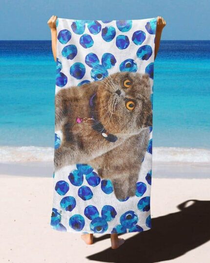 A person stands barefoot on a sandy beach, holding up the Custom Design 3D Printed Beach Towel – Blueberry, which features a gray cat and blue polka dots, with turquoise ocean water and a blue sky in the background.