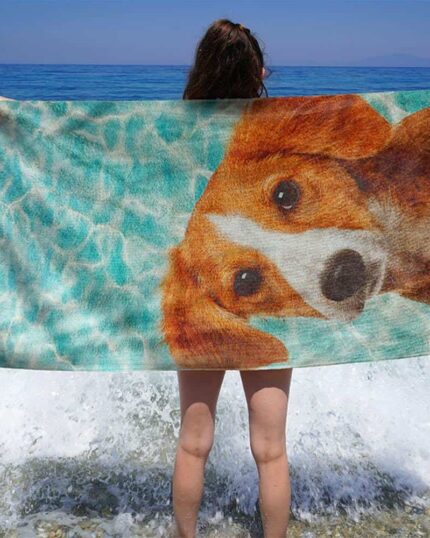 A person stands on a rocky beach facing the sea, holding up a Custom Design 3D Printed Beach Towel – Pineapple. Waves crash around their legs as they display the vibrant towel featuring a realistic pineapple design against the blue water.
