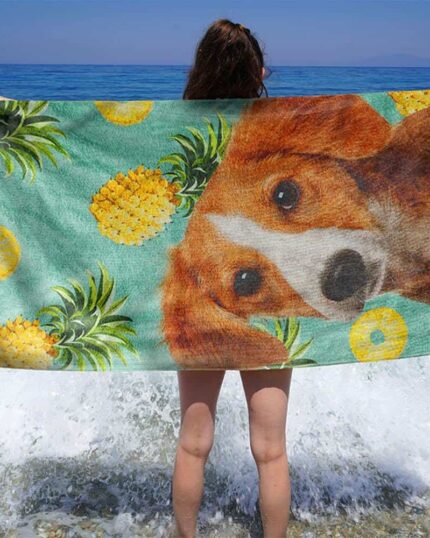 Bir kişi kıyıda durmuş, büyük bir köpek yüzü ve ananasların resmedildiği evcil hayvan tuval sanatından esinlenerek tasarlanan Özel Tasarım 3D Baskılı Plaj Havlusu – Ananas'ı tutuyor. Dalgalar ayaklarının dibinde kırılırken, arkasında mavi deniz uzanıyor.