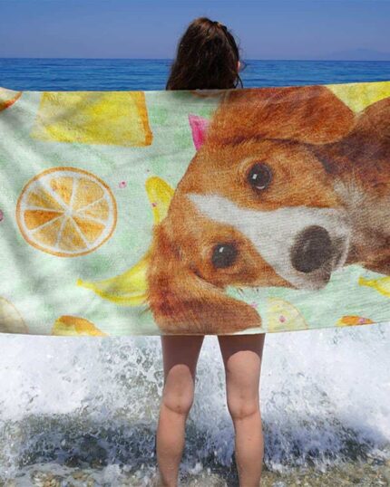 A person stands on the beach with waves crashing, holding a Custom Design 3D Printed Beach Towel – Pineapple, featuring a brown and white dog among colorful fruit illustrations like lemon slices, bananas, and pineapples.