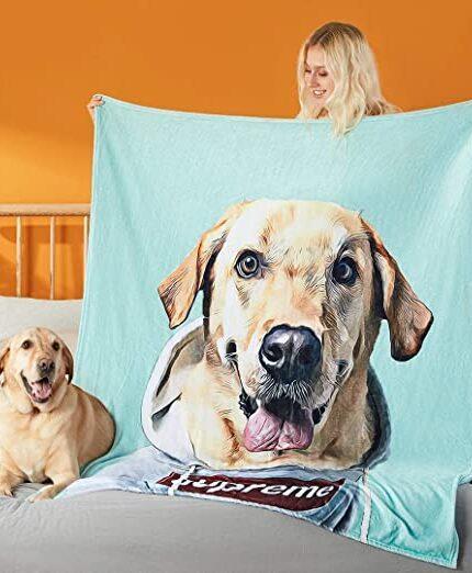 A woman holds up a light blue Custom Design Pop-Art Blanket – Solid Colors with a large, realistic Labrador in a hoodie, while a similar Labrador lies nearby on the bed against an orange wall.