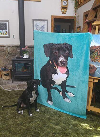 A black and white dog sits on a green carpet next to a large turquoise Custom Design Pop-Art Blanket – Solid Colors featuring the dog's image. The rustic room has wooden accents and a wood stove.