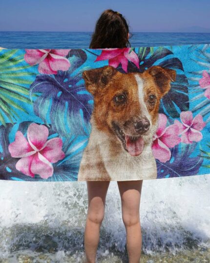 Okyanusun kenarında duran bir kişi, pembe çiçekler ve yemyeşil tropikal yapraklarla süslenmiş bir köpek resminin yer aldığı Custom Design 3D Printed Beach Towel – Tropical Leaves plaj havlusunu tutuyor. Dalgalar ayaklarına sıçrarken.