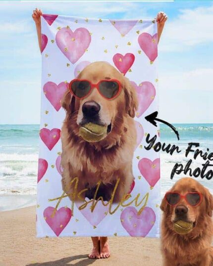 A person holds a Personalised 3D Printed Beach Towel with a custom pet design of a dog in red sunglasses and a ball, pink hearts around. The real dog, also with red sunglasses, sits on the sand by the ocean. Text: “Ashley” and “Your friends photo.”.