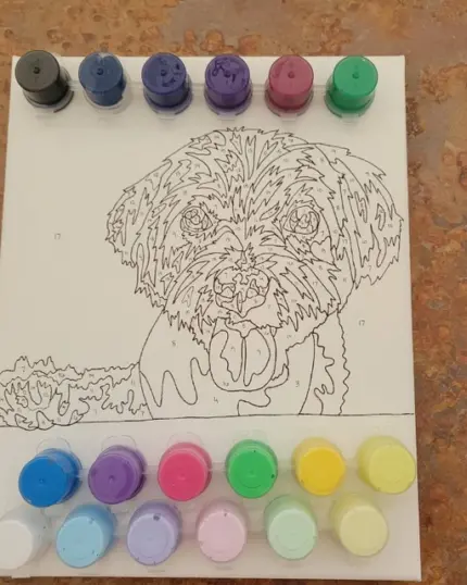 The Handmade Paint by Number Kit features a custom dog outline with its tongue out and includes small assorted paint pots arranged around the canvas on a brown, textured surface.