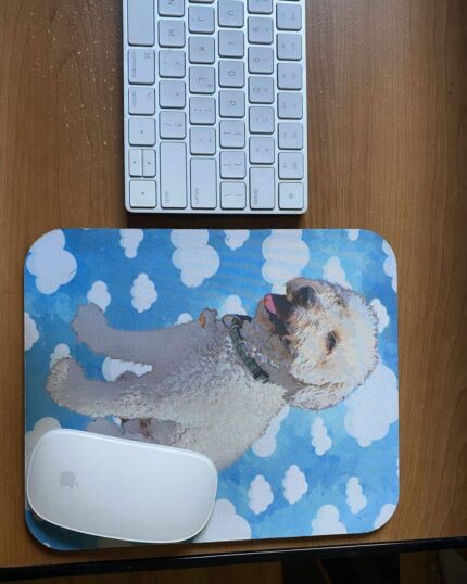 A white keyboard and mouse rest on a wooden desk. The mouse is on a blue Custom Pet Art Mouse Pad with clouds and a happy, fluffy white dog, highlighting the charm of personalized pet-themed designs.