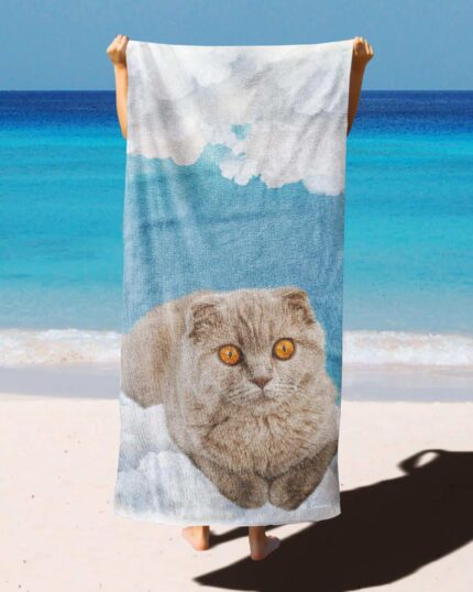 On a sandy beach with the ocean and sky behind, a person holds up the Custom Design 3D Printed Beach Towel – Clouds, featuring a gray cat with orange eyes lying on clouds.