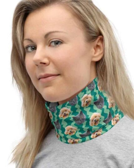 A light-haired woman, dressed in a light gray top and smiling gently, models the Custom Neck Gaiter With Pet Faces featuring teal fabric printed with cats and dogs, standing against a plain background.