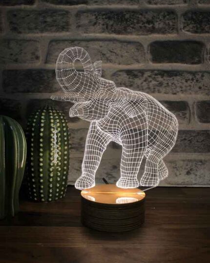 The 3D Elephant LED Night Lamp emits a white glow on a wooden table next to a cactus ornament and pet canvas, all set against a brick wall background.
