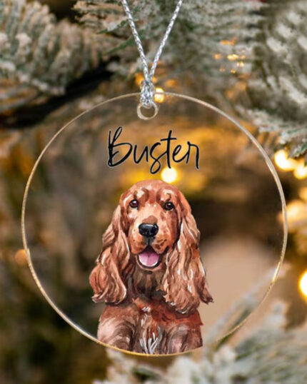 The Personalized Transparent Pet Ornament hangs on a Christmas tree, displaying a painted brown dog with long ears and the name Bugten. Warm lights and pine needles glow in the background, capturing custom pet art charm.