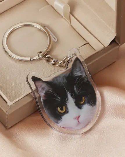 The Personalized Animal Printed Plexiglass Keychain features a clear acrylic charm with a black and white cat face, displayed beside an open beige gift box on satin—ideal for custom pet accessory fans.