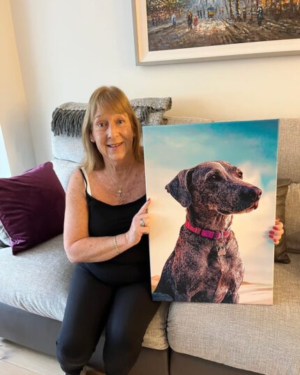 A smiling woman with light brown hair sits on a sofa, holding a vibrant Custom Design Abstract Pet Portrait of a dog in a pink collar. The portrait shows the dog against a bright backdrop, with more artwork hanging behind her.