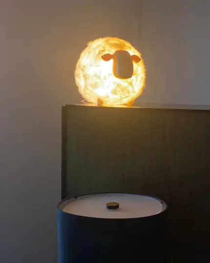 The Soft Cotton Sheep Night Light, shaped like a sheep's head, sits on a wooden cabinet above a light gray cylinder—an ideal addition to neutral indoor spaces for showcasing custom design pet canvas artwork.