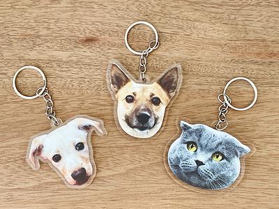 Three Personalized Animal Printed Plexiglass Keychains—two dogs and a gray cat with cartoon faces on a wooden surface—ideal for pet lovers and fans of creative custom design.