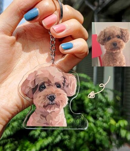 A hand with colorful nails holds a Personalized Animal Printed Plexiglass Keychain featuring an illustrated brown, curly-haired dog matching the photo inset. Greenery and a building appear in the background. Ideal for custom pet art lovers.