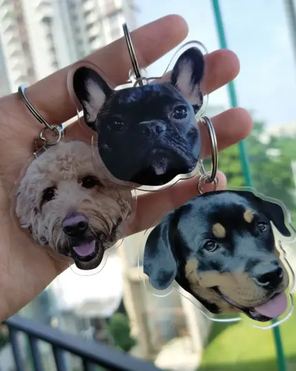 A hand holds three Personalized Animal Printed Plexiglass Keychains with cutout dog faces—a curly-haired light dog, a black French bulldog, and a black-and-tan dog—highlighting custom pet art against a city backdrop.